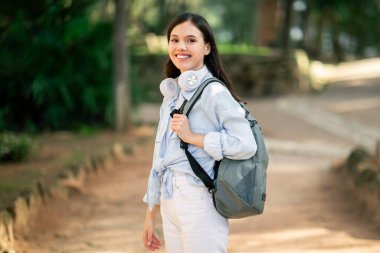 Günlük kıyafetli genç bir kadın kulaklıklarını omuzunda taşıyor ve halk parkında yürürken sıcak bir şekilde gülümsüyor.