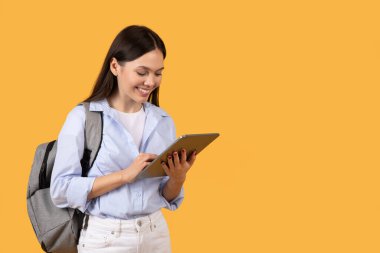 Dijital tablet kullanan odaklanmış genç bir kadın, sarı arka planda sırt çantasıyla gelişigüzel giyinmiş.