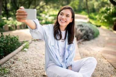 Excited young woman captures a selfie in a park, reflecting the trend of self-documentation and social media clipart