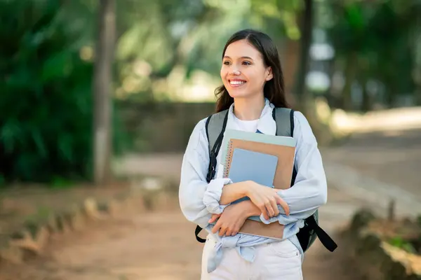笑顔の若い女性は 公共の公園 コピースペースで屋外に立っているようにノートとバックパックを保持しています — ストック写真