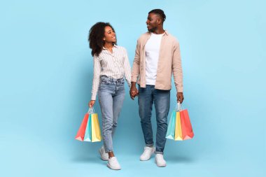 Mutlu bir Afro-Amerikan çifti mavi bir arka planda yürürken ve alışveriş torbaları taşırken birbirine bakar.