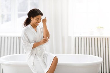 Beyaz bornozlu Afro-Amerikan kadın temiz, parlak bir banyoda, küvetin kenarına oturmuş, vücut kremi kullanıyor.