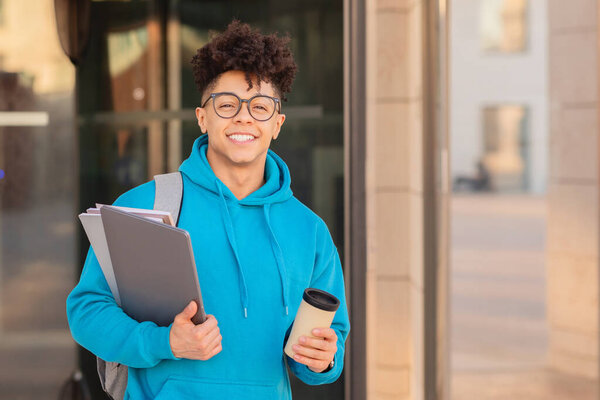 Радостный студент-бразилец с рюкзаком и чашкой кофе, в очках и синей толстовке в городской обстановке