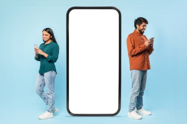 İki neşeli genç Hintli çift akıllı telefonlarına odaklanmış büyük boş bir akıllı telefon ekranının yanında maket için duruyorlar.
