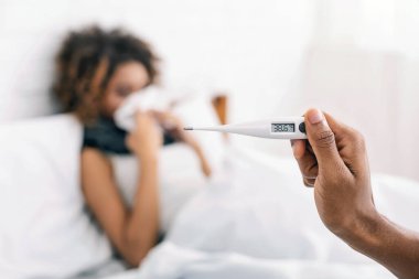 Ateşli hasta Afro-Amerikalı kadın yatakta yatıyor, termometreyi 38.6 C ile tutan adam.