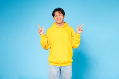 A joyful Asian teenage boy in a yellow hoodie posing with peace signs against a vivid blue studio background clipart