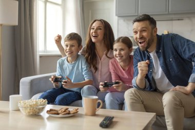 Heyecanlı bir aile babası, anne ve çocuklar keyifle ve kahkahalarla kanepede oyun oynuyorlar.