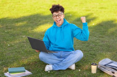 Elinde dizüstü bilgisayarla çimenlerde oturan neşeli Brezilyalı bir öğrenci başarısını, defterlerini ve kahve fincanını kutluyor.