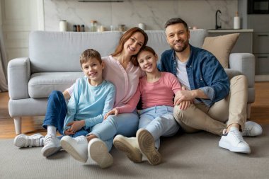 Gülümseyen aile babası ve annesi ve iki çocuğu çağdaş bir oturma odasındaki kanepede uzanıyorlar.