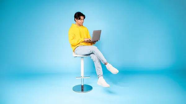 十几岁的亚洲男孩坐在高凳子上 用笔记本电脑 在蓝色的背景下带着沉思的表情 — 图库照片
