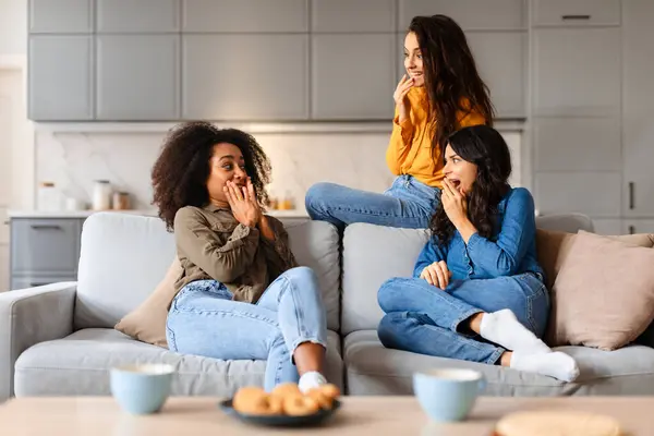 stock image A candid moment captures three diverse young women sharing laughter while sitting comfortably on a light grey couch in a cozy living room setting
