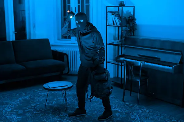 A mysterious figure with a flashlight explores a room bathed in blue light, the atmosphere is tense and intriguing