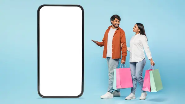 ヒンドゥーのカップルは 青い背景に対してモバイルショッピングを意味する大規模な空白の画面スマートフォンの横にショッピングバッグを保持しています — ストック写真