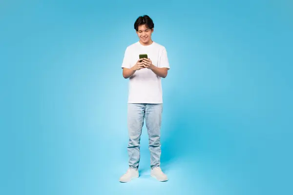 若いティーンアジアの男深く彼のスマートフォンを使用して魅了 技術エンゲージメントを示す 青い背景 — ストック写真