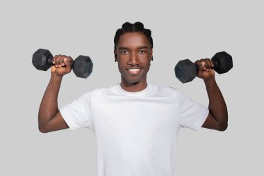 A fit and happy young African American man stands lifting dumbbells while smiling confidently against a neutral backdrop clipart