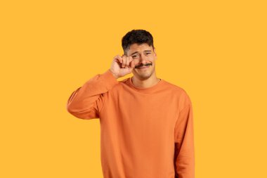 Tearful emotional man with moustache in an orange sweater wiping away a tear on a yellow background clipart