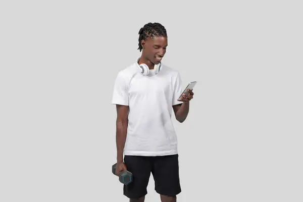 スポーツアフリカ系アメリカ人男性は 白い背景にダンベルを保持しながらスマートフォン画面を見ています — ストック写真