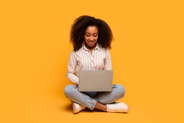 Dizüstü bilgisayarı üzerinde çalışırken yerde bağdaş kurup gülümseyen Afro-Amerikan kadın.