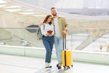 Mutlu çift havaalanı terminalinde sarı bir bavulla yürürken kucaklaşıyor, neşeli eşler pasaportlarını tutuyor ve kameraya bakıyor, birlikte seyahat etmekten zevk alıyor, uzayı kopyalıyor.