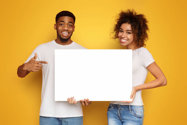 Young happy african-american couple holding blank advertising board, orange background, copy space