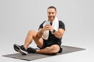 Cheerful man using a smartphone while sitting on a fitness mat in an exercise studio setting, nice app clipart