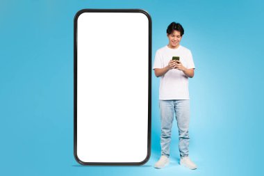 Genç bir Asyalı, reklam içeriği için ideal boş bir ekranla büyük bir telefon ekranının yanında duruyor.