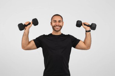 Formda olan adam kameraya gülümserken, beyaz arka planda spor kıyafetleriyle rahatça ağırlık kaldırıyor.