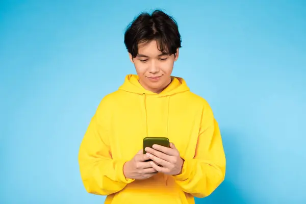 青い背景に携帯電話にテキストを送ることに深く関わった黄色い汗着のアジアの若い男 — ストック写真