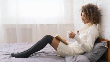 Kitap okuyan ve yatakta kahve içen genç bir kadın.