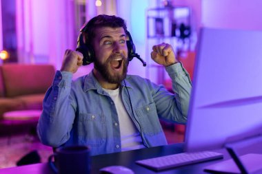 Kulaklıklı neşeli bir adam yumruklarını kaldırır zaferle bilgisayar masasında otururken, renkli ortam ışıklarıyla aydınlanır.