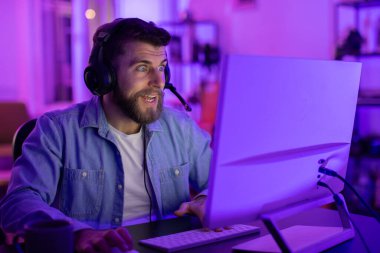 Kulaklıklı adam yoğun bir şekilde bilgisayar ekranına odaklanmış, evdeki kasvetli mavi ışıklı oda ile çevrili.