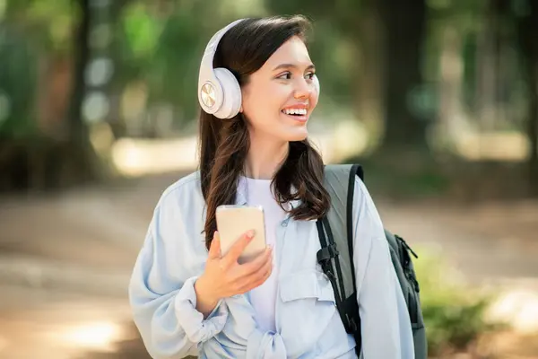 年轻的快乐的欧洲姑娘 戴着耳机 一边用智能手机欣赏着音乐 一边带着柔和的微笑向远方望去 一边走在外面一个安静的公园里 关门了 — 图库照片