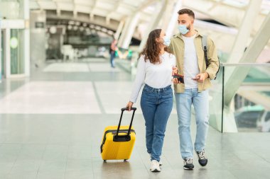 Yüz maskesi takan genç bir çift modern bir havaalanında bavullarıyla yürüyor.