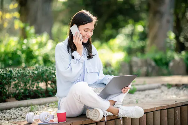 Multitasking european young woman in a relaxed outdoor setting, cheerfully talking on the phone while working on a laptop, with a coffee cup and headphones nearby, outside