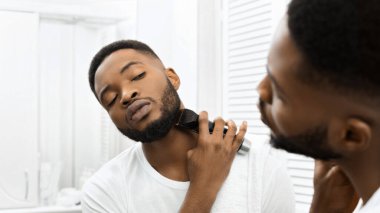 Odaklanmış Afrikalı Amerikalı adam sakal bırakıyor. İyi ışıklandırılmış, modern bir banyo ortamında budayıcı kullanıyor.