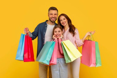 Mutlu bir aile gururla renkli alışveriş çantalarını sergiliyor, neşe ve aile birliğini sarı bir arka planda sergiliyor.