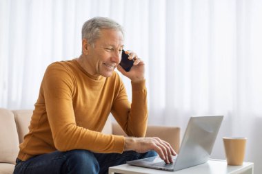 Yaşlı bir adam, rahat bir oturma odasında dizüstü bilgisayarıyla birden fazla iş yaparken bir telefon görüşmesi yapar.