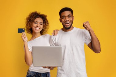 Gülümseyen Afrikalı genç erkek ve kadın ellerinde dizüstü bilgisayar ve kredi kartı, online alışveriş başarısı veya kolay elektronik işlemleri gösteriyor