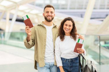 Neşeli erkek ve kadın mutlu yüzler sergiliyorlar, ellerinde pasaportlar ve uçuş kartları var, modern bir havaalanı ortamında.