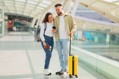 Akıllı telefonlu bir ulaşım merkezinde yürüyen gülümseyen bir çift, bağlantıyı resmediyor ve modern seyahat