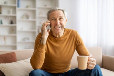 Neşeli, yaşlı bir adam elinde kahve fincanıyla akıllı bir telefonla sohbet ediyor. İyi aydınlatılmış bir oturma odasında oturuyor.