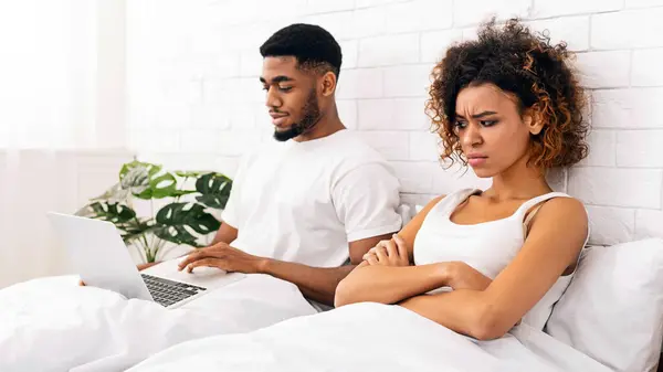 Technology and relationship problems. Addicted young black man lying in bed with laptop, woman looking angry and offended, empty space
