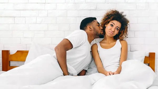 An affectionate scene where African American young man kisses a woman on the cheek in a cozy bedroom setting