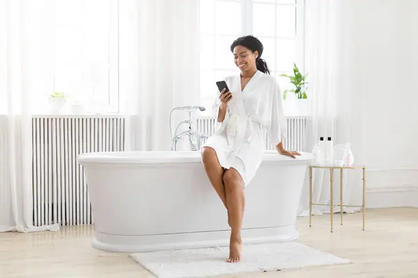 非洲裔美国女人在明亮的浴室里享受着轻松的时光 她在浴缸边使用智能手机 — 图库照片