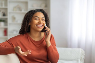 Kanepede oturmuş gülümseyen siyah bir kadın cep telefonuyla konuşuyor, mutlu bir Afrikalı Amerikalı kadın evde cep telefonuyla konuşuyor.