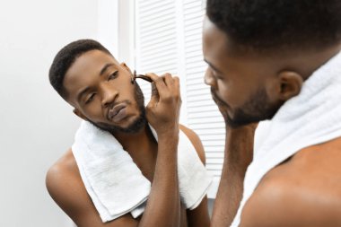 A young African american man with a towel around his neck looks at his reflection in the mirror, shaving beard clipart