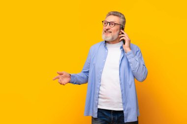 Sarı arka planda cep telefonuyla konuşan, gözlüklü, neşeli, yaşlı, gri saçlı adam cihaz aracılığıyla iletişim kuruyor. Mobil iletişim kavramı. Metin için boş alan