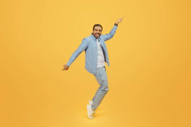 An exuberant bearded indian man dances with joy, his arm outstretched pointing upwards, wearing a denim shirt and glasses, against a yellow backdrop clipart