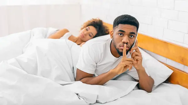 African American man looks worried during a phone call as his partner naps, reflecting relationship and communication themes
