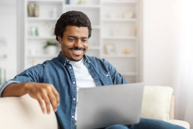 Evdeki kanepede rahat bir şekilde otururken dizüstü bilgisayarıyla meşgul olan neşeli siyah bir adam..
